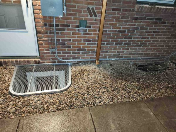 Brick building exterior with window well covered by metal grate, gravel ground.