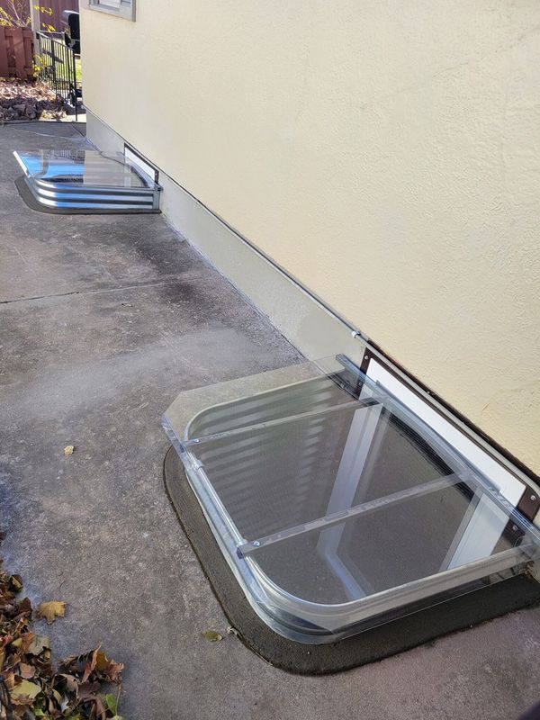 Two basement window wells with clear covers set into concrete next to a light-colored building.