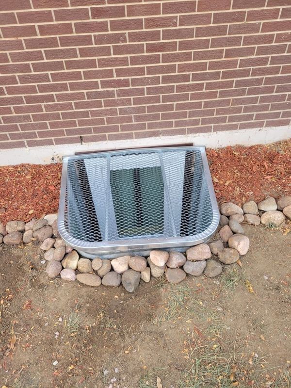 Window well with metal grate, bordered by rocks and reddish mulch, against a brick wall.