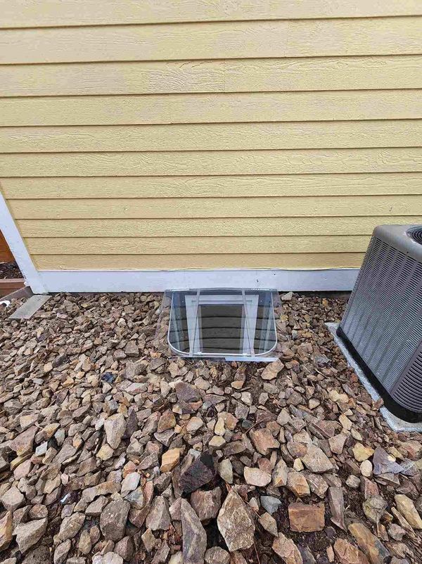 Window well with grate below yellow siding, surrounded by gravel.