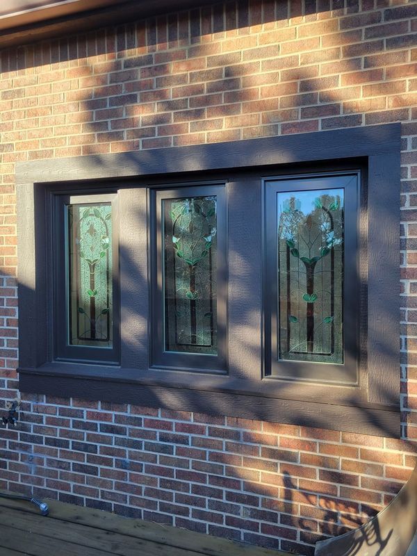 Three stained glass windows in a dark brown frame set against a brick wall.