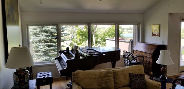 A living room with a grand piano by a window, a sofa, and other furniture. The view outside is a landscape.