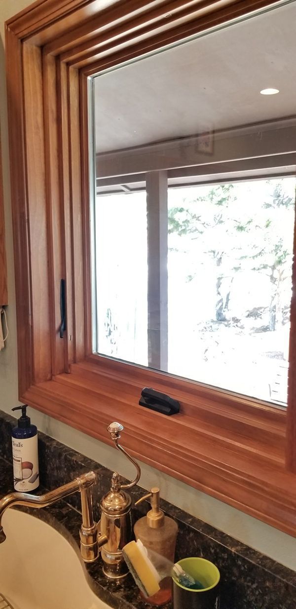 Bathroom window with brown wood frame, looking out to greenery. Sink and faucet in foreground.