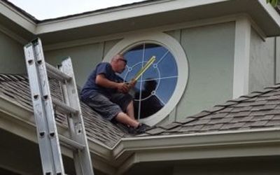 Man on roof near round window, using tool. Ladder beside him.
