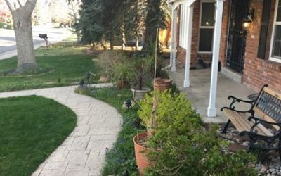 Brick house with a winding walkway, green lawn, trees, and potted plants on a porch with a bench.