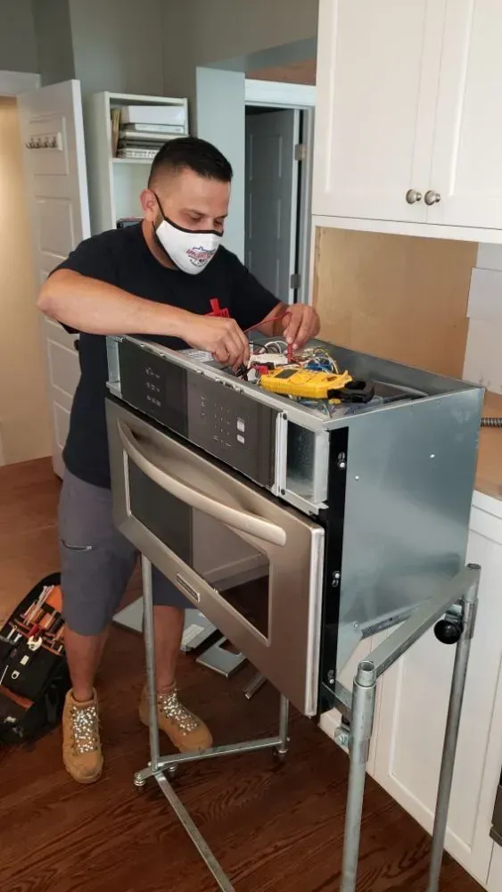 Man in mask installing an oven in a kitchen, standing on wood floor. Cabinets and tools are visible.