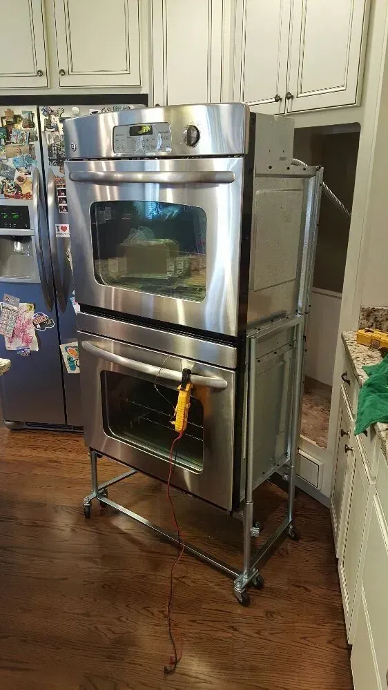 Stainless steel double oven on a metal cart in a kitchen, resting on wheels.