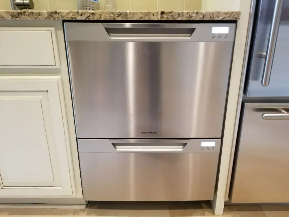 Stainless steel, two-drawer dishwasher installed in a kitchen. Cabinets on either side, granite countertop above.