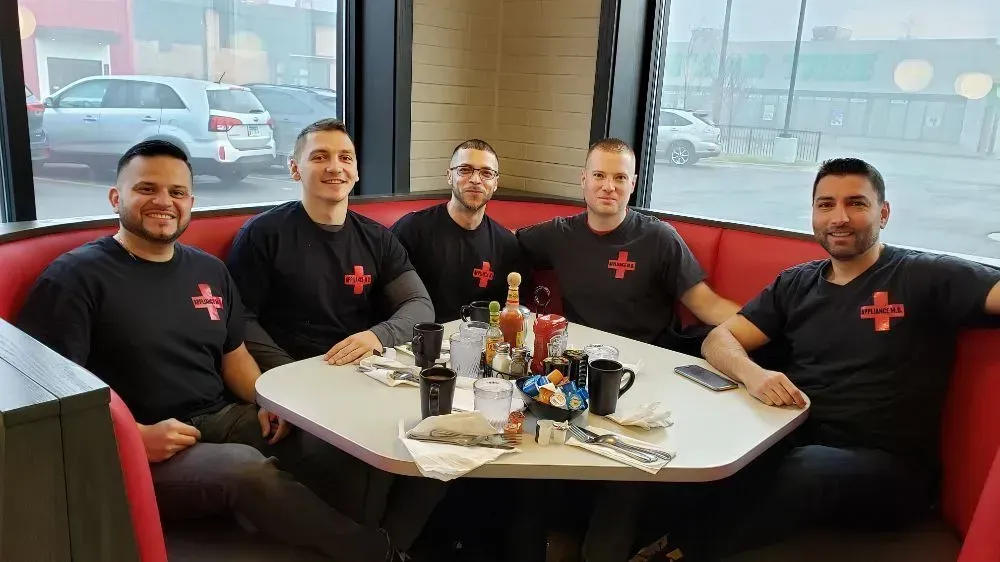 Five men in black t-shirts with red crosses sit around a table in a diner booth, smiling.