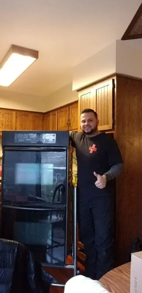 A man stands beside a double oven in a kitchen, giving a thumbs up. He has a beard and wears a black shirt.
