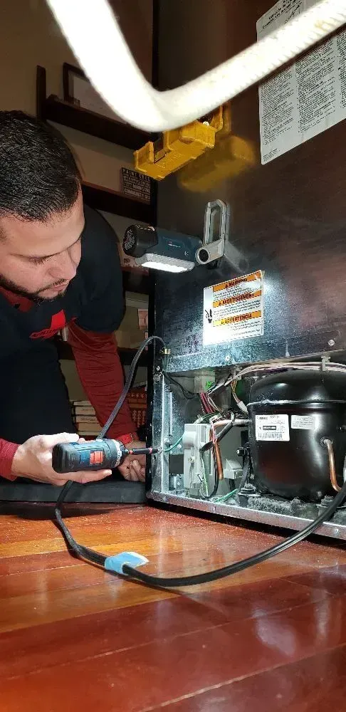 A man repairs a refrigerator, using a screwdriver. Black and red clothing and a brown floor.
