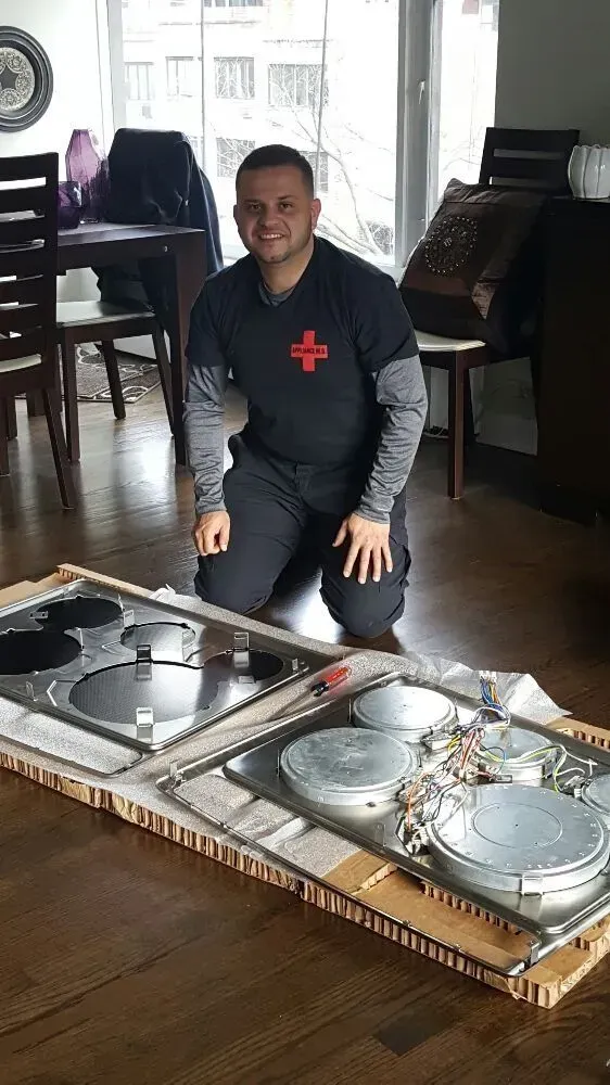 Man kneeling by a dismantled stove, smiling. Indoors, near a window. Black shirt with red cross.
