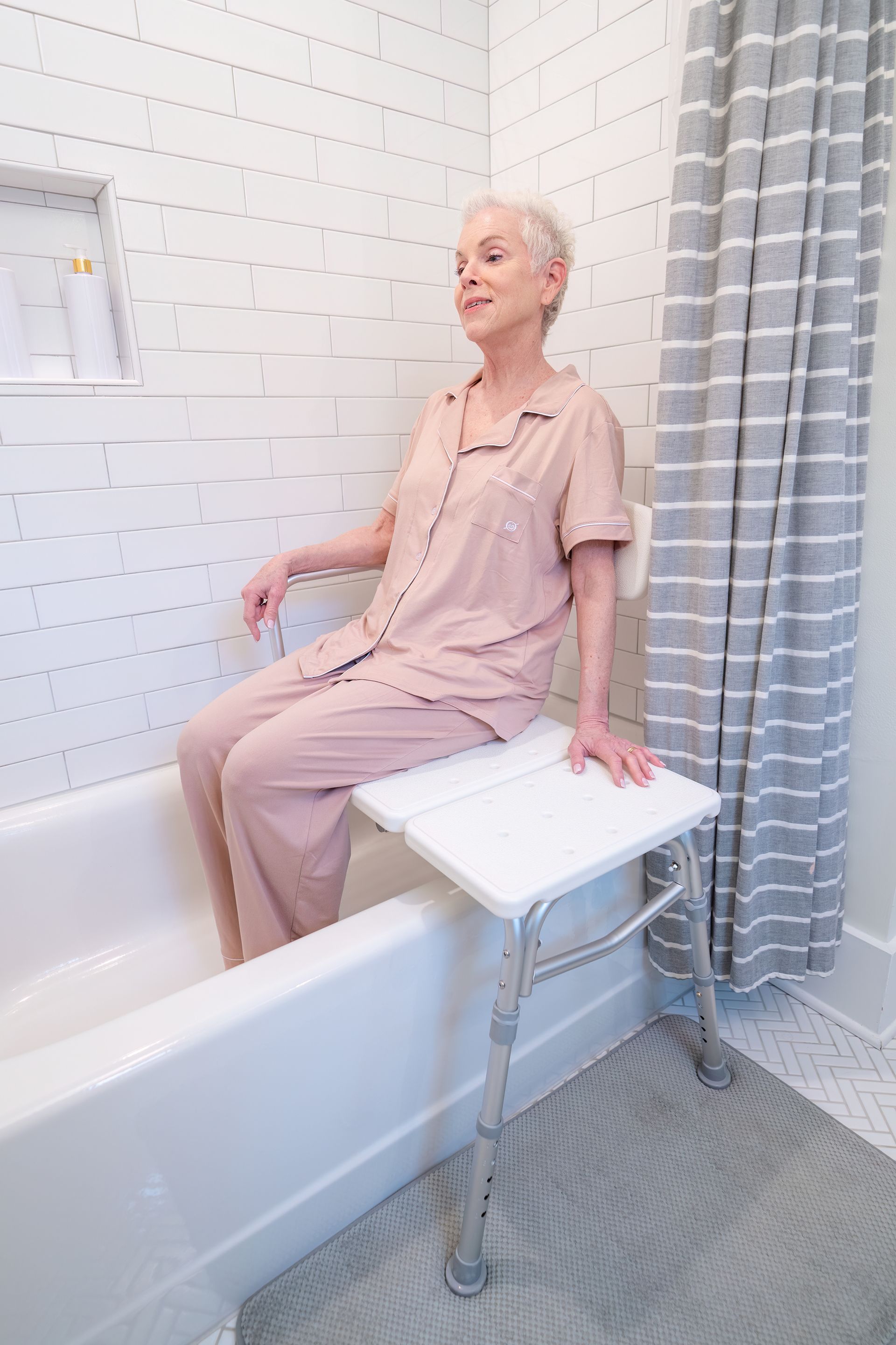 A person sitting on a white bathtub transfer bench inside a tiled shower with a gray striped curtain.