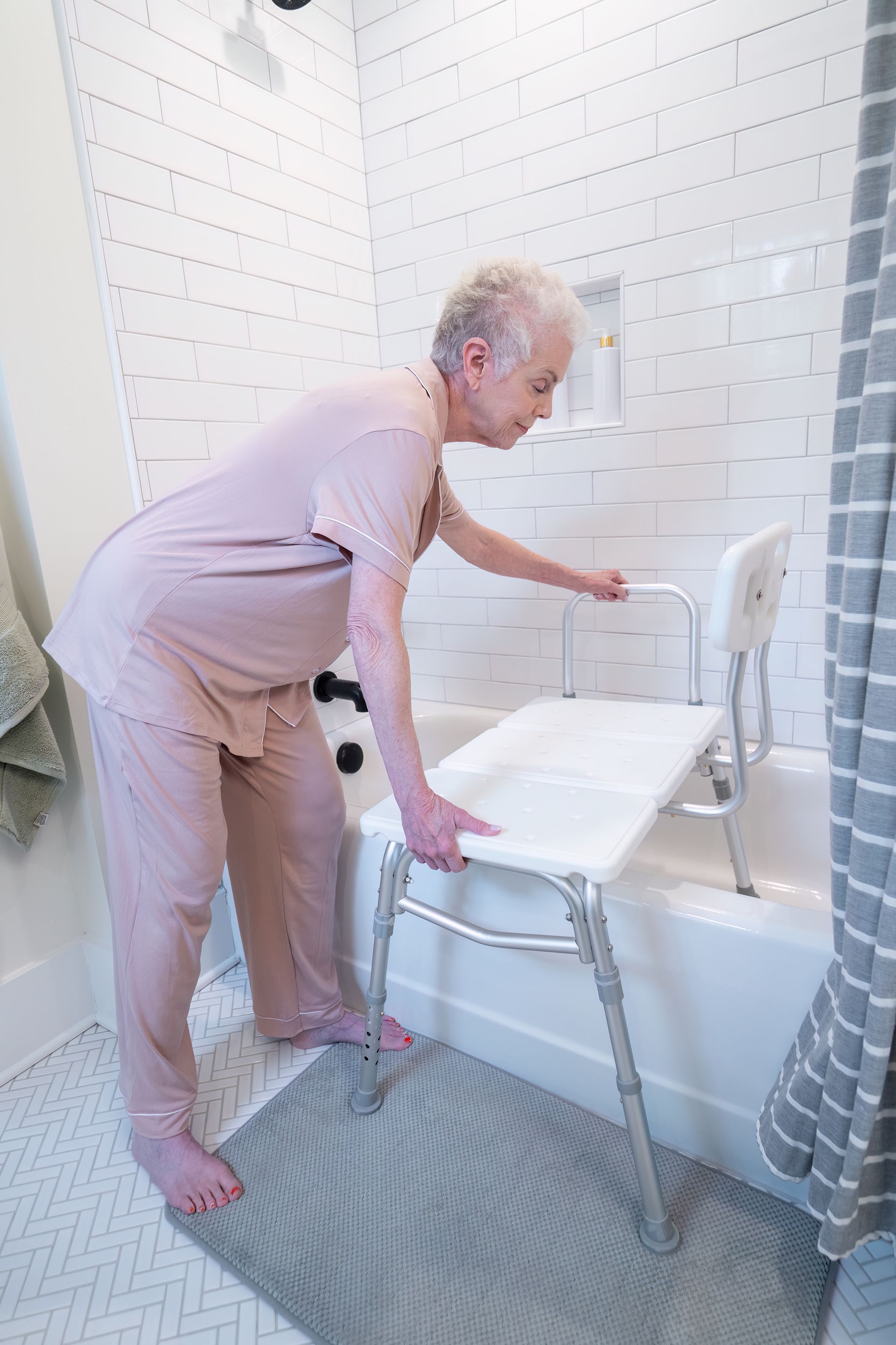 A person using a white tub transfer bench with a backrest to safely enter a shower with white tile walls.