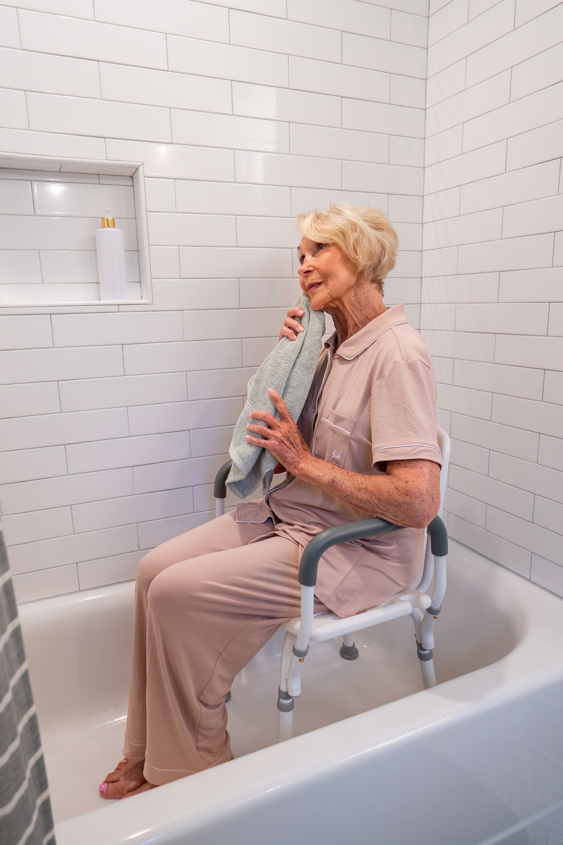 A person sitting on a white shower chair inside a bathtub, holding a towel to their face in a tiled bathroom.