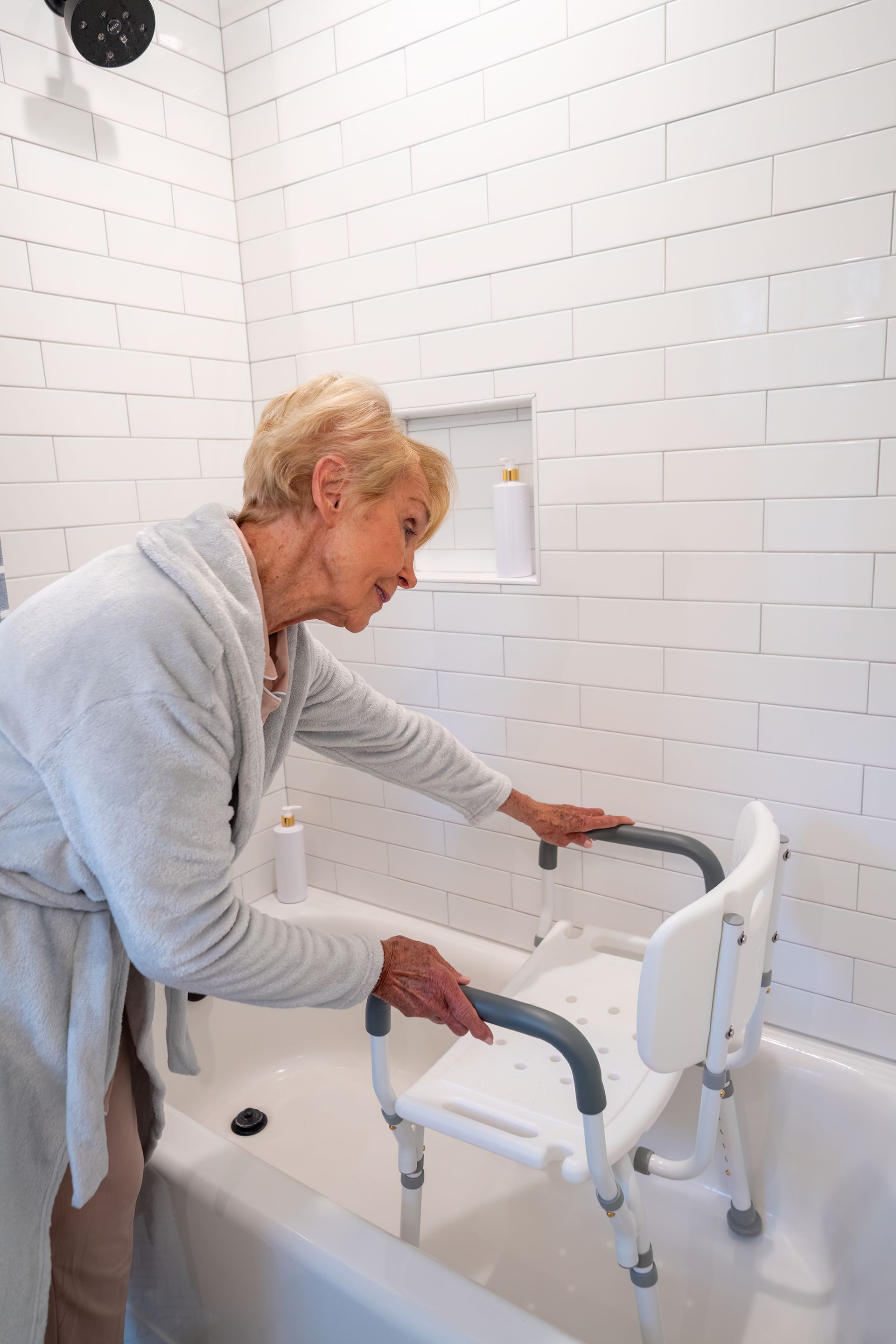 An elderly person in a bathrobe reaching to adjust a white shower chair installed inside a white bathtub.