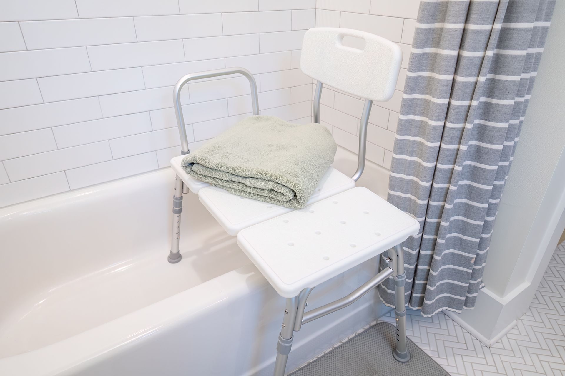 A white bathtub transfer bench with an armrest and backrest sits over the edge of a tub against a white tiled wall.