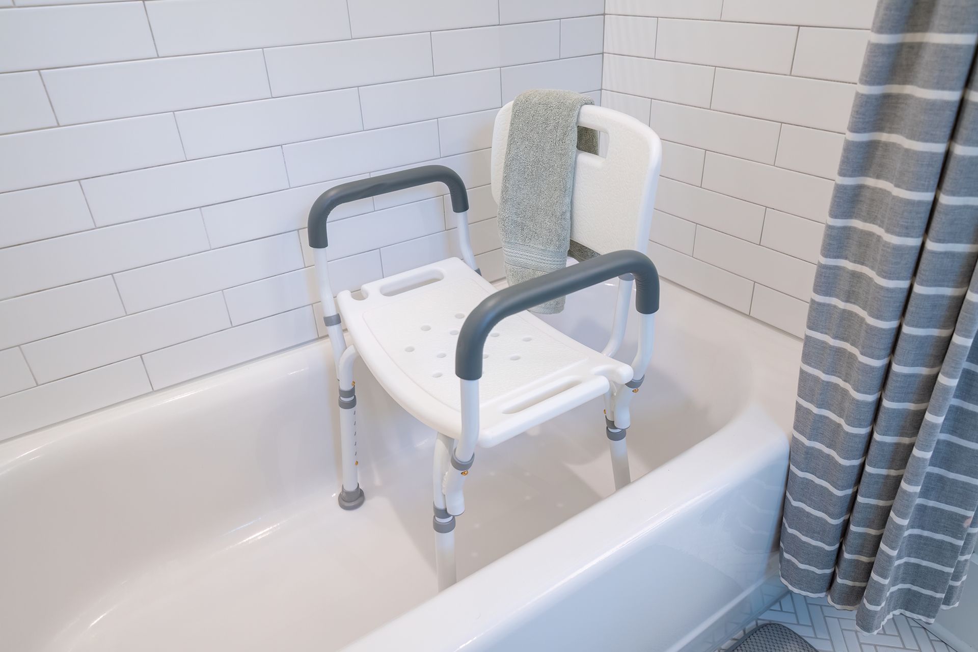 A white shower chair with grey armrests and a backrest sits inside a bathtub against a white tiled wall.