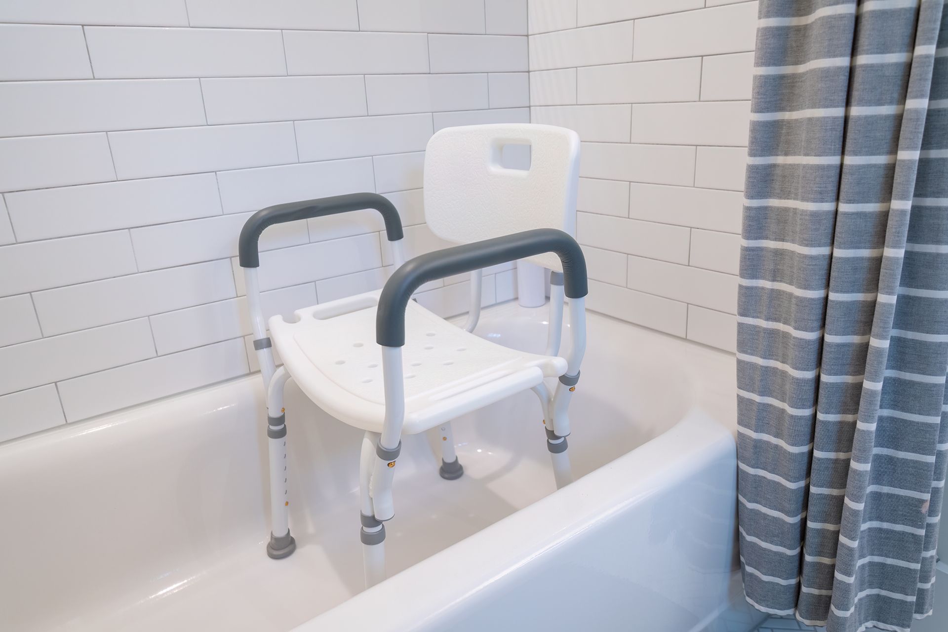 A white shower chair with dark gray armrests sits inside a bathtub against a white tiled wall and a striped shower curtain.
