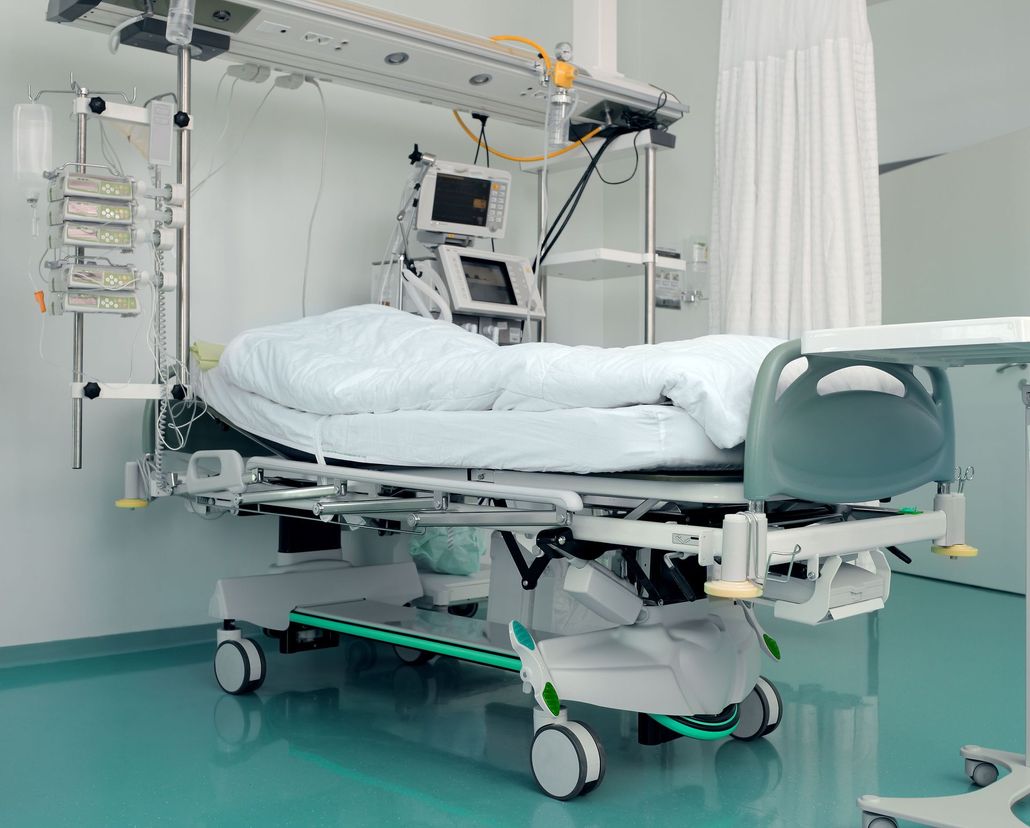 An empty hospital bed with medical equipment and monitors positioned above and behind it in a clinical room.