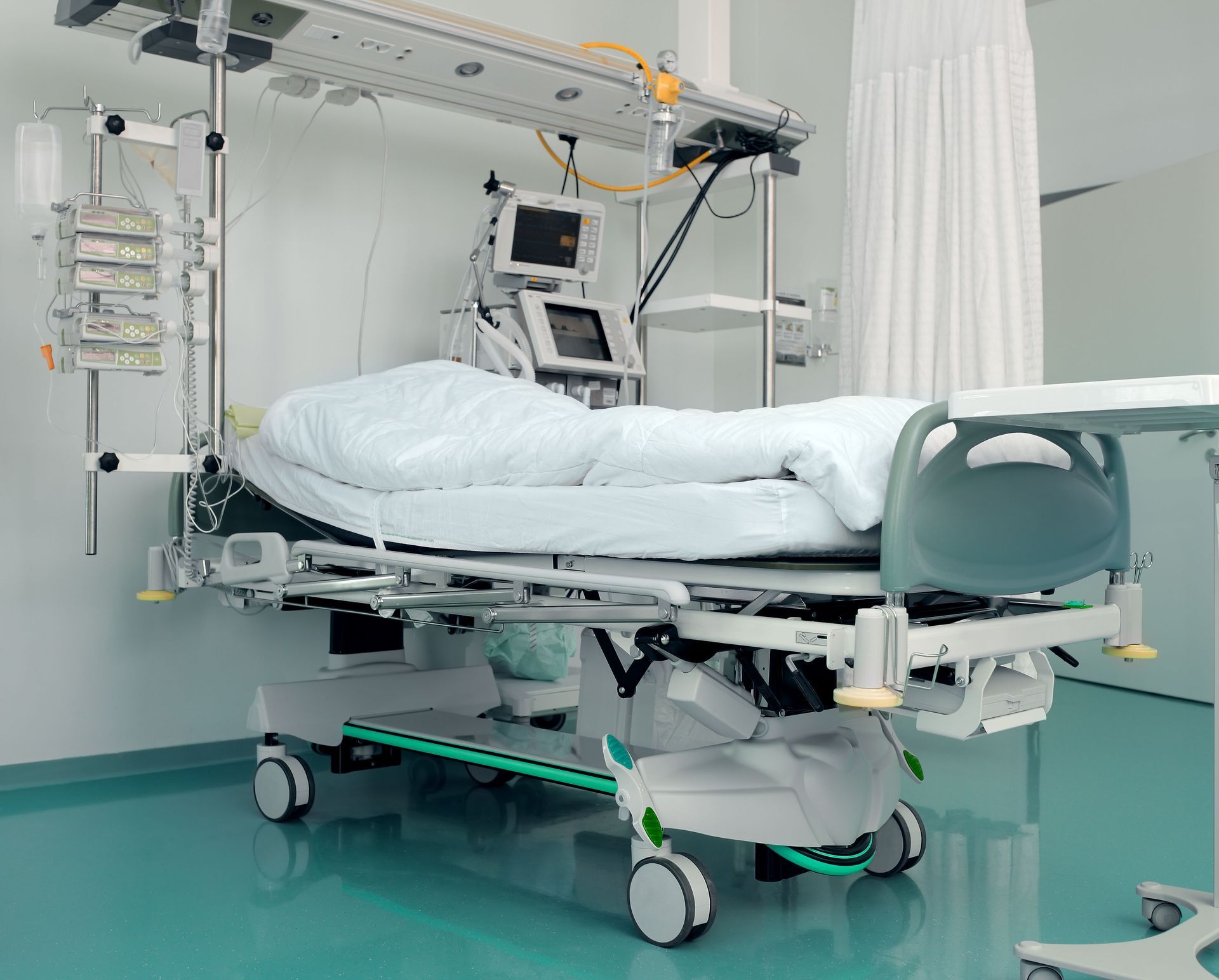 An empty hospital bed with medical equipment and monitors positioned above and behind it in a clinical room.