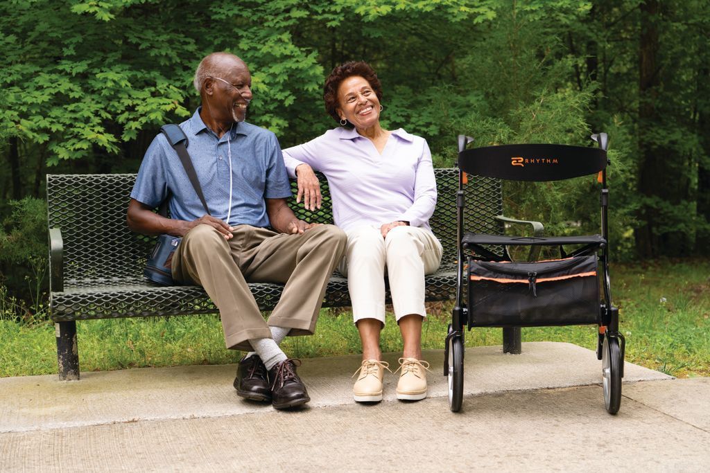 Two people sit on a park bench next to a rolling mobility walker, smiling and looking upward in a natural setting.