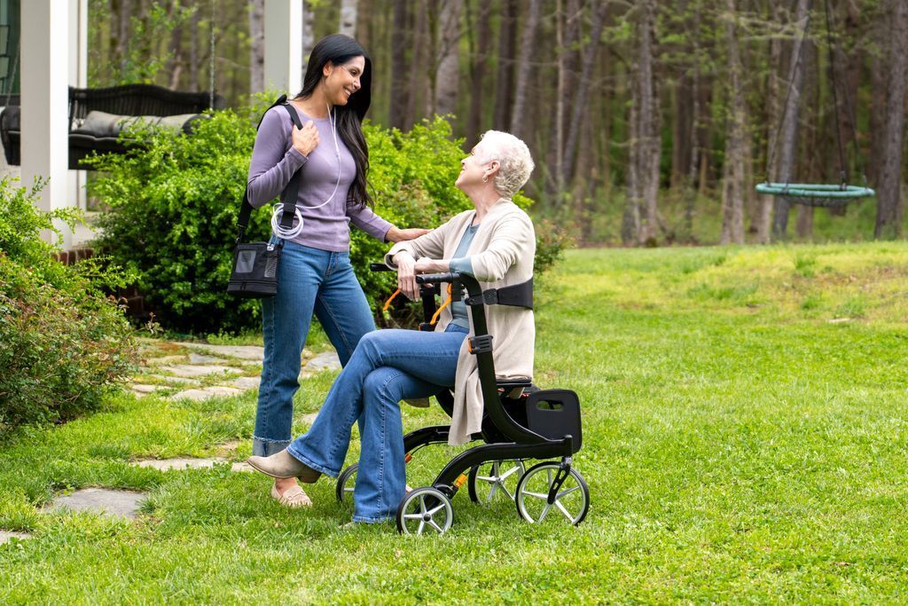Two people interact while one sits on a rollator walker in a grassy yard near a house.