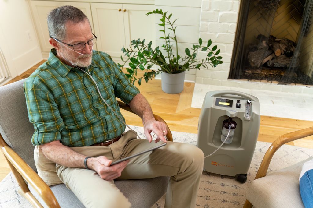 A man in a green shirt sits in a chair using a tablet with a nasal cannula connected to an oxygen concentrator nearby.
