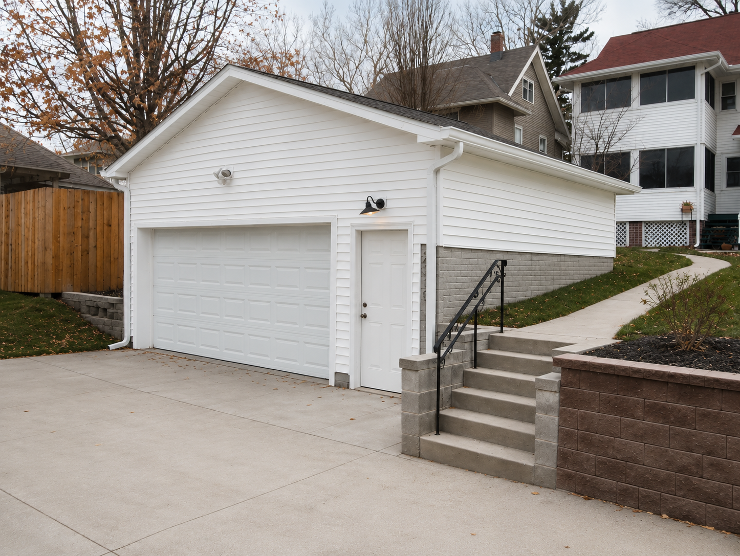 Detached white garage with attached side door, concrete driveway, and stairway beside a retaining wall