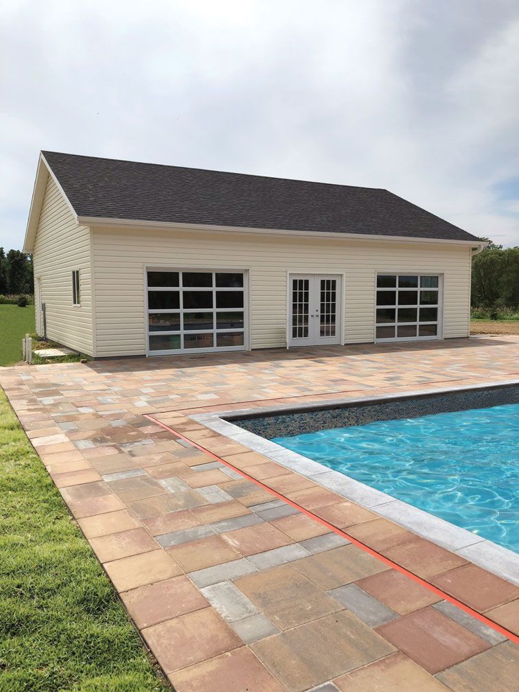 Poolside building with glass doors and windows, light siding, dark roof, brick patio, and pool.