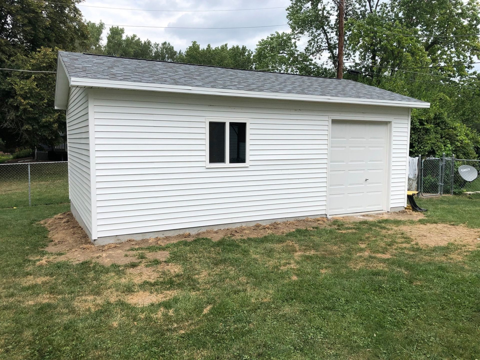 White shed with gray roof and a single window and garage door, on a grassy lot.
