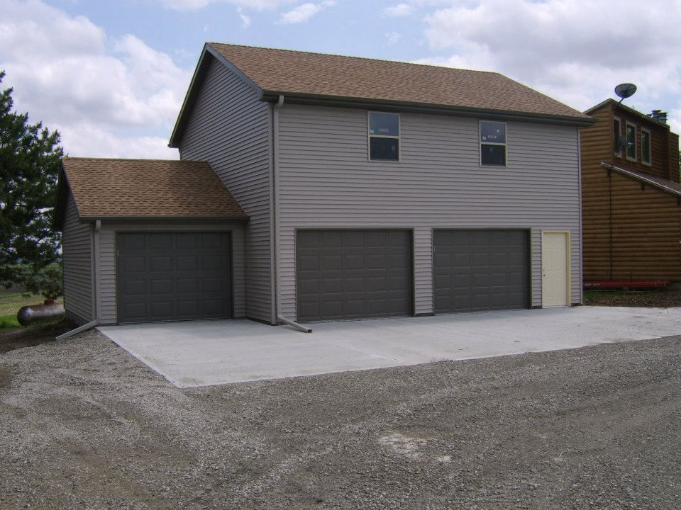 Two-story garage with three bays and brown siding. Concrete driveway, gravel parking area, and brown roof.