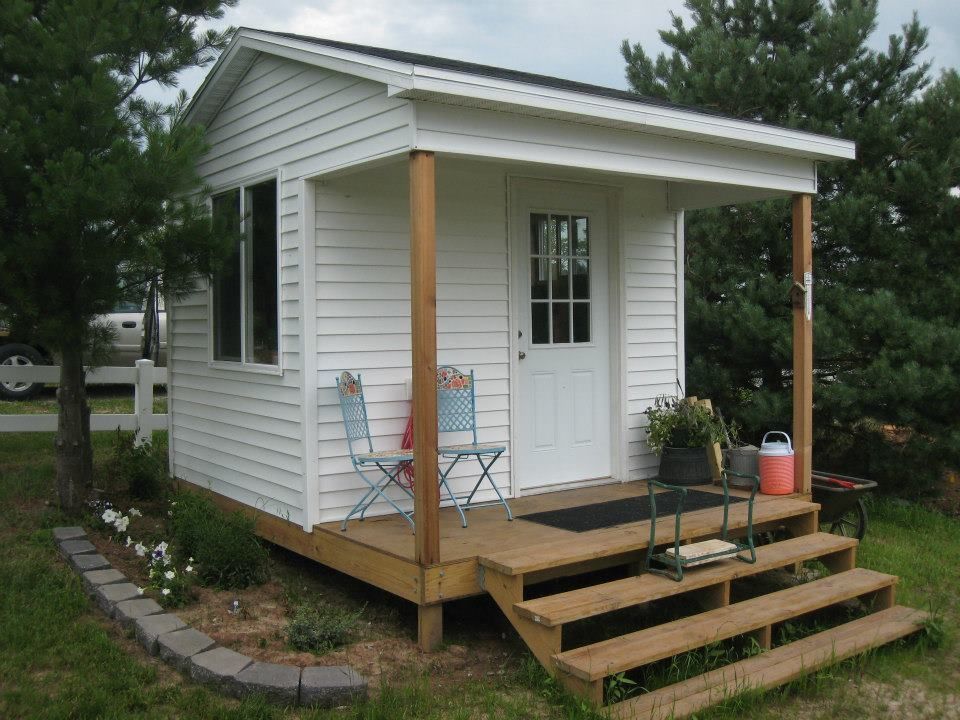 Small white shed with porch, wooden steps, and two blue chairs.