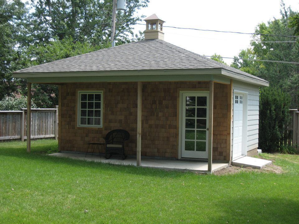 Small wood-shingled building with a porch, a window, and a door on a grassy lawn.