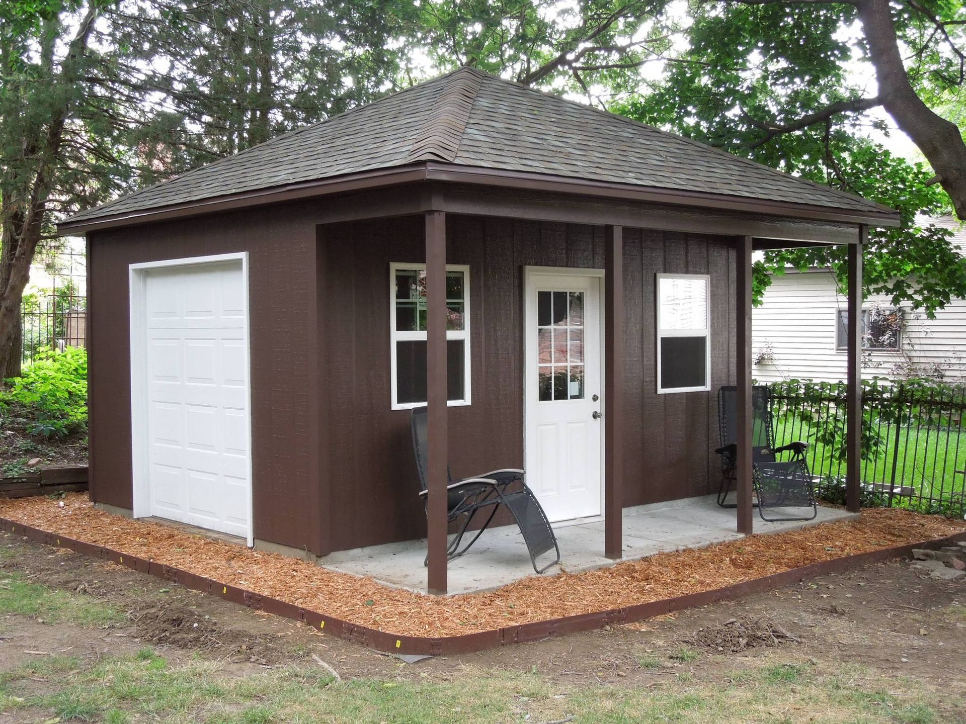 Brown shed with white garage door and porch, surrounded by mulch, and small lawn chairs.