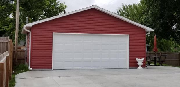 Red-sided garage with a white door and concrete driveway. A white urn and outdoor furniture are to the right.