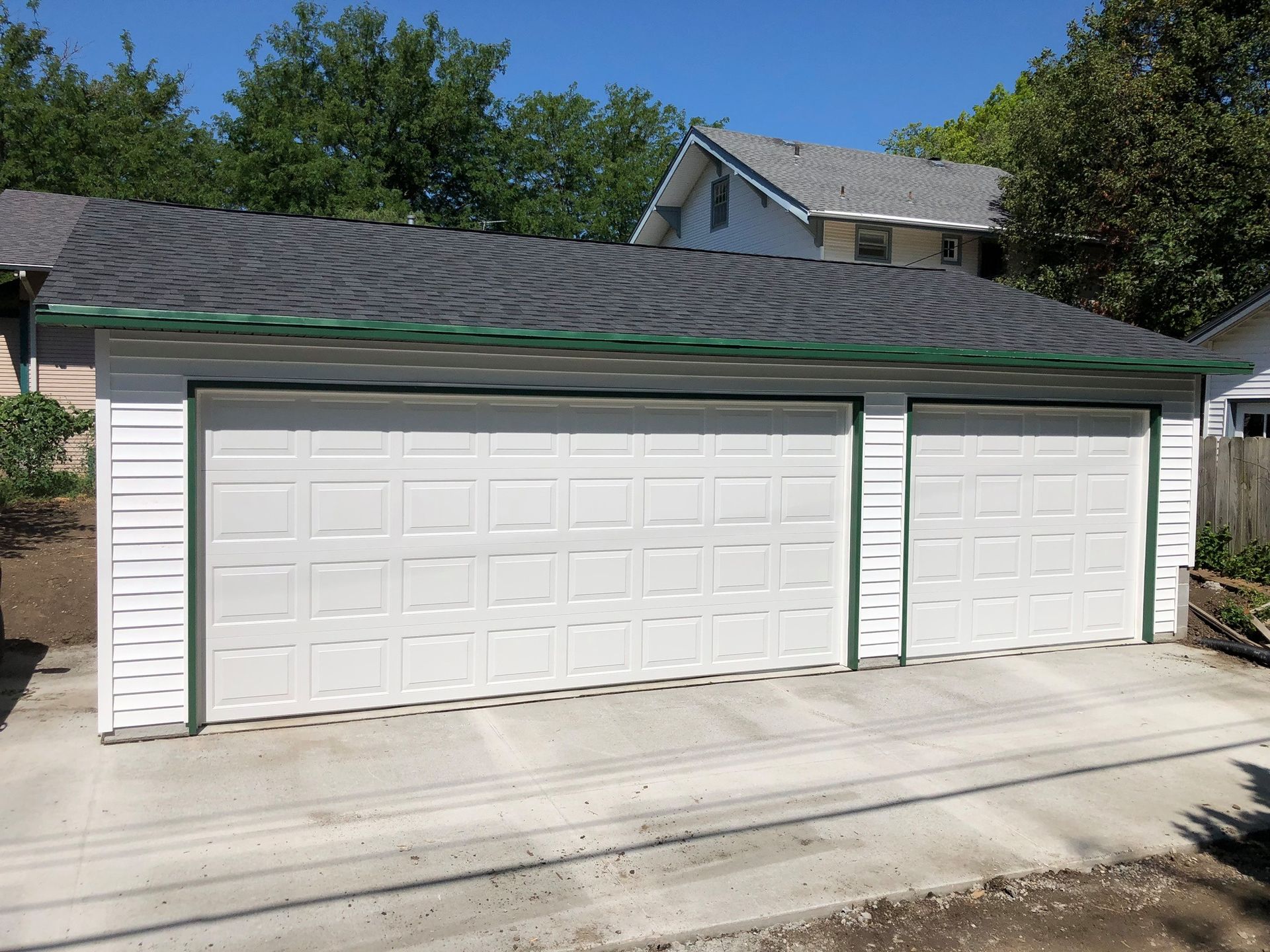 Two-car white garage with green trim, dark roof, concrete driveway, and trees in background.