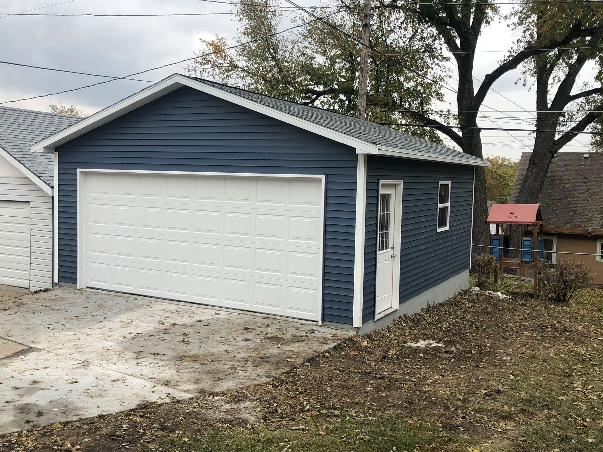 Blue-sided detached garage with white door and garage door; gray roof; in front yard.