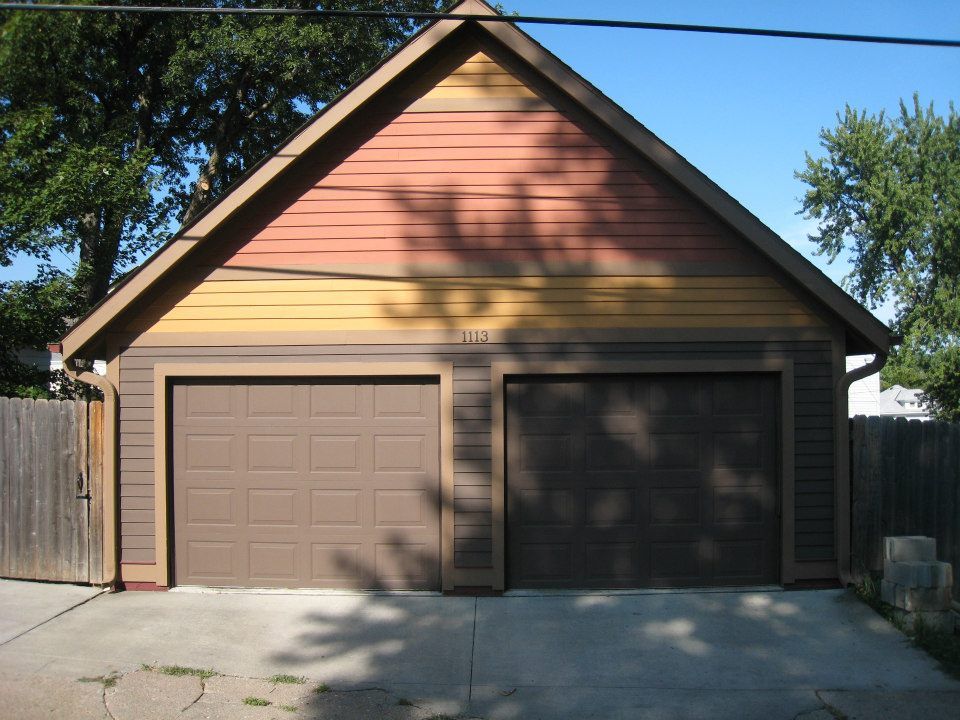 Two-car garage with brown doors and colorful siding. Trees and fence in the background.