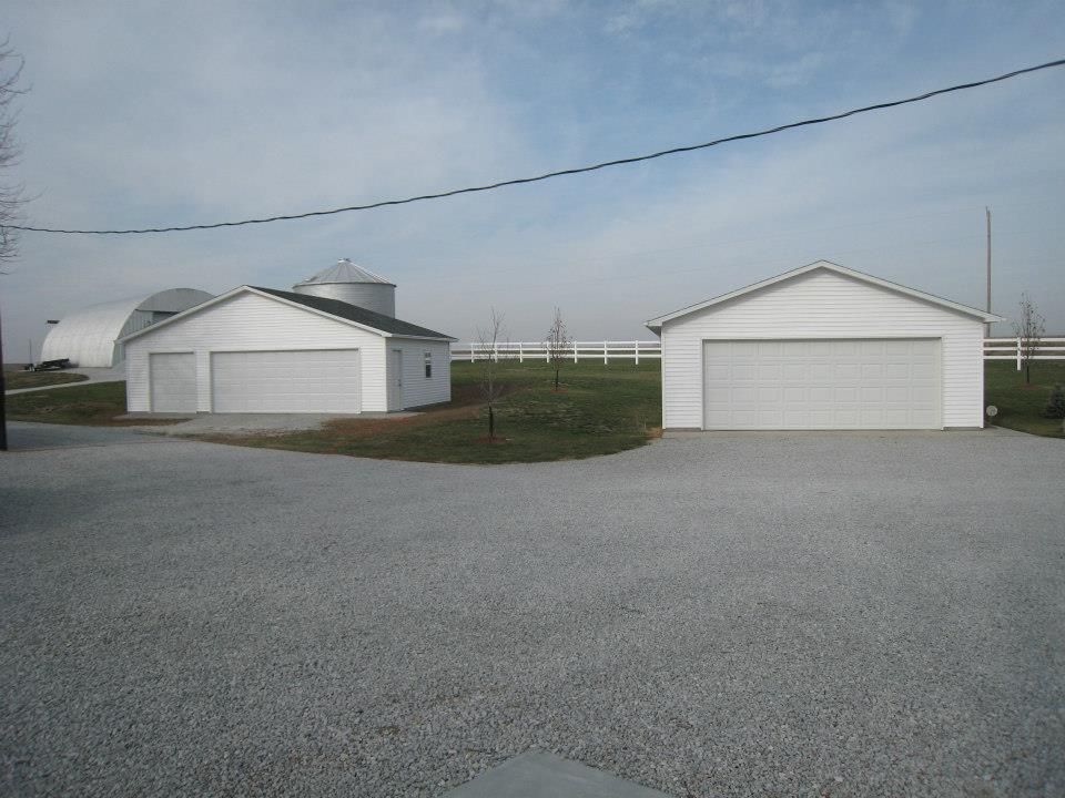 Two white garages on a gravel drive, with a farm building and silo in the background under a cloudy sky.