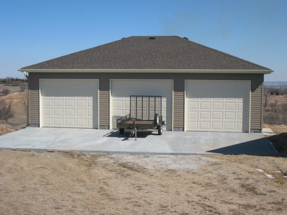 Three-car garage with beige doors, brown roof, and a trailer parked in front on a concrete slab.