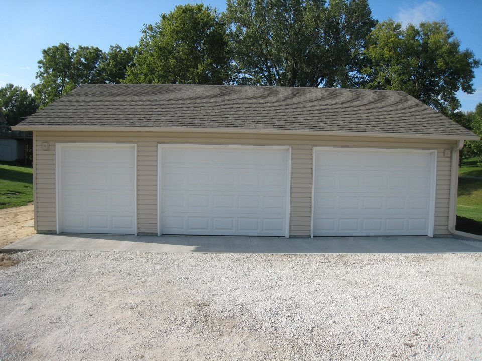 Three-car beige garage with white doors, gravel driveway, and trees in the background under a blue sky.