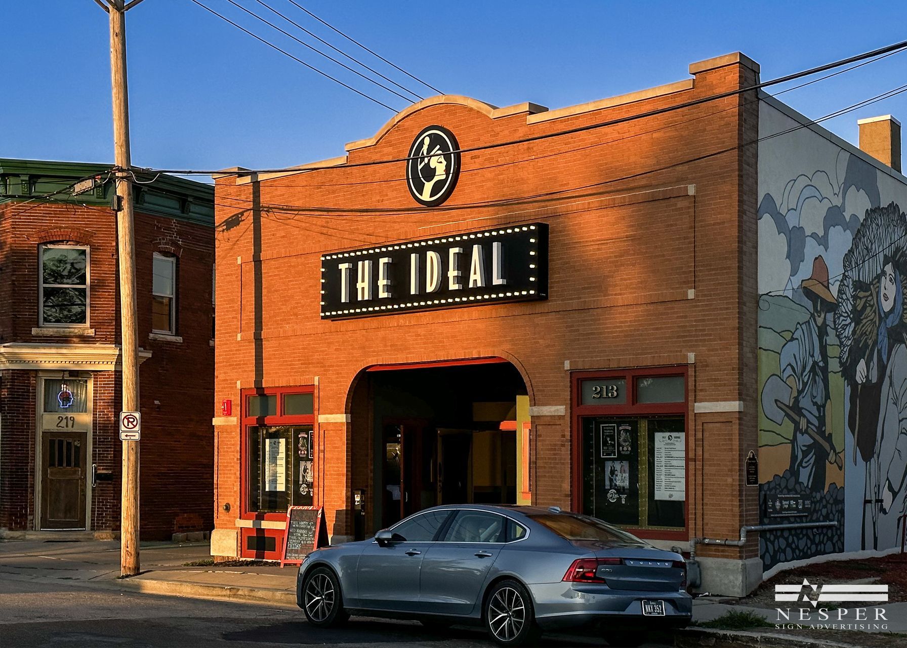 The Ideal Bar in red brick building with sign, street view, car parked. Mural on side.