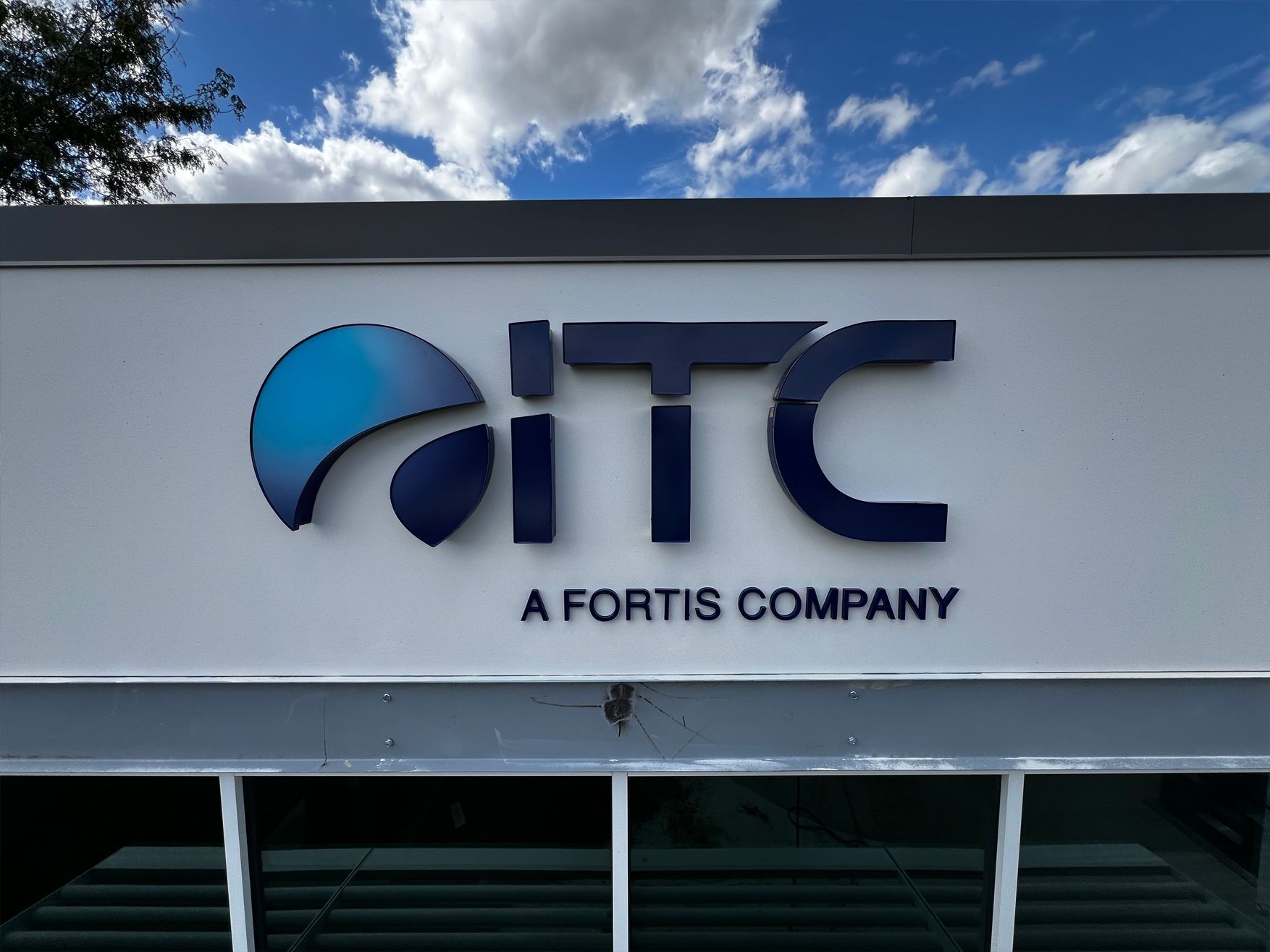 Sign for ITC, a Fortis company, with blue logo against a white background, under a partly cloudy sky.