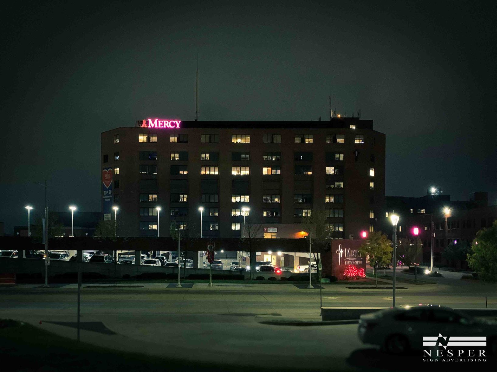Nighttime view of Mercy Hospital building with illuminated 'Mercy' sign. Cars and streetlights in the foreground.
