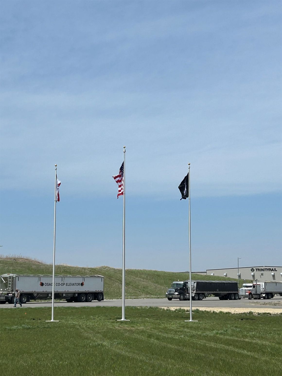 Three flag poles with flags and three semi trucks on a grassy area under a blue sky.