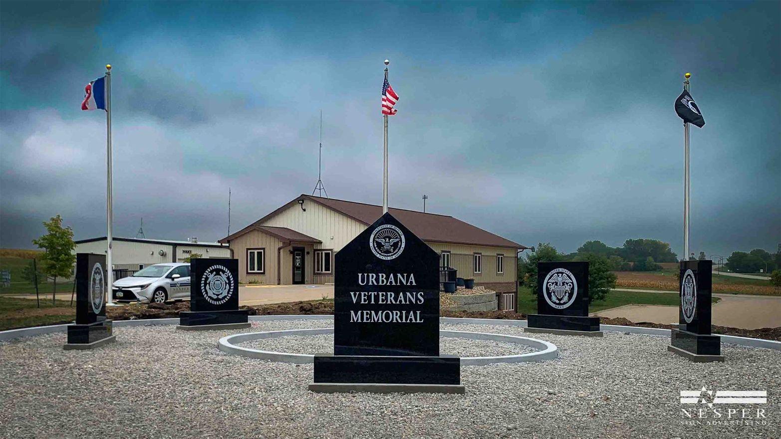 Veterans memorial with US flag and other flags; black stone monuments in a circle. Building in the background.