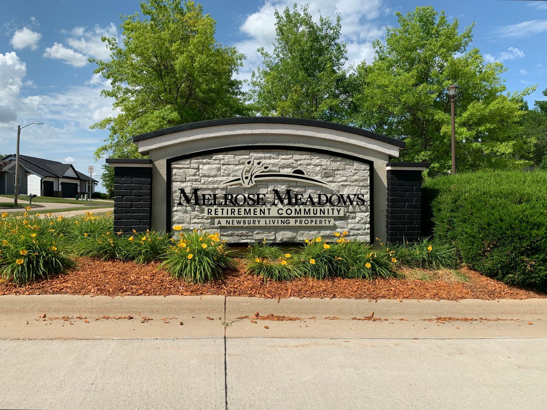 Sign for Melrose Meadows housing community. Stone facade, arched top, bordered by landscaping and trees.