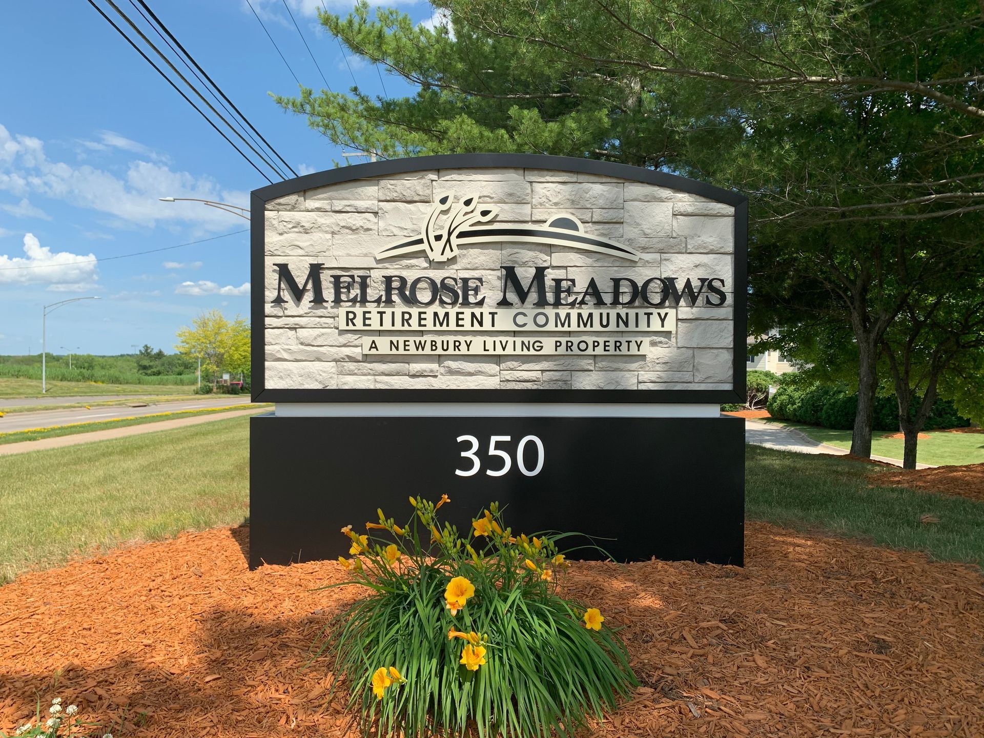 Sign for Melrose Meadows Retirement Community, 350 address; black and stone-look panels, yellow flowers, blue sky.