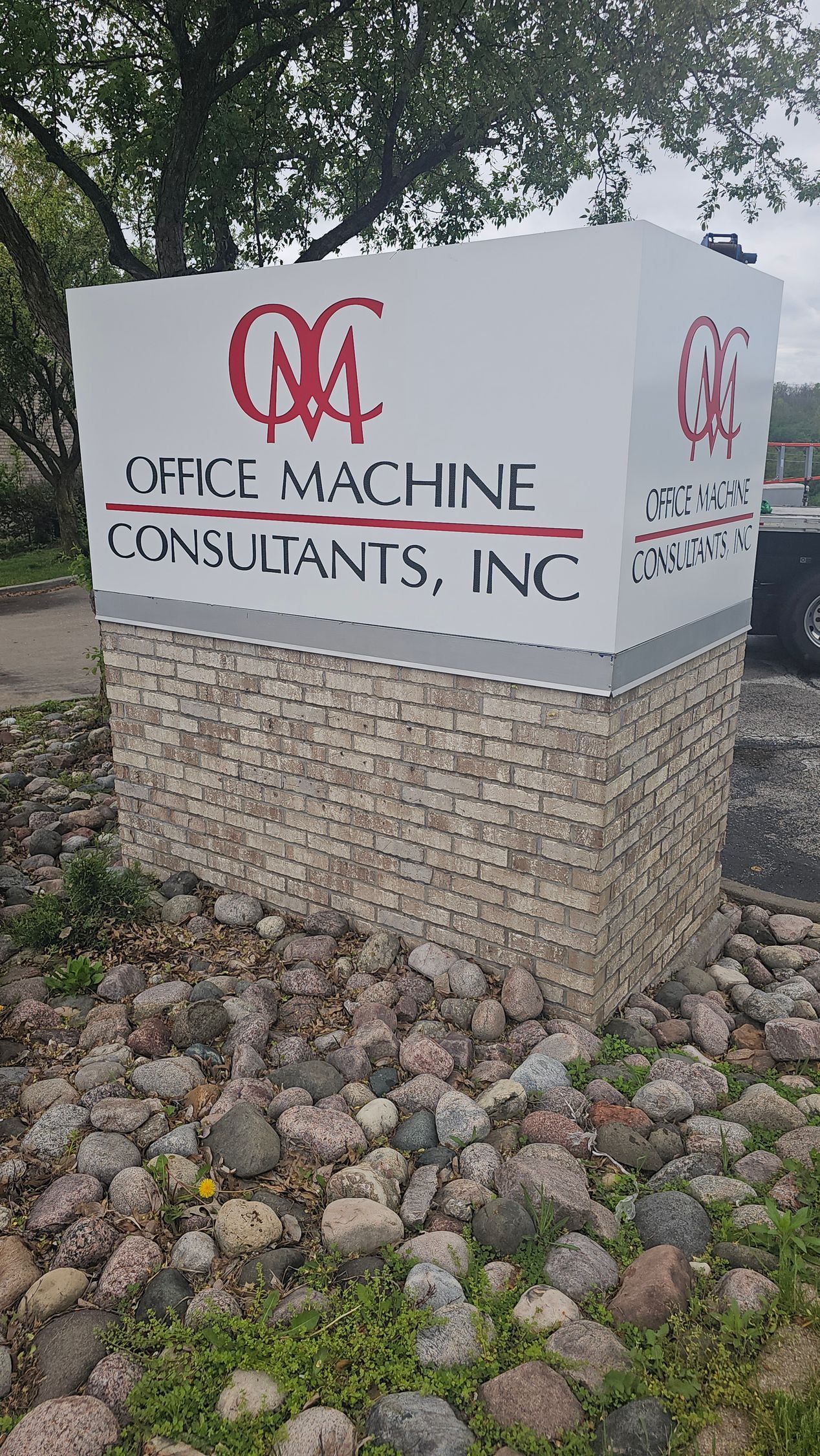 Sign for Office Machine Consultants, Inc., with red logo on white, supported by stone-covered column, surrounded by rocks.